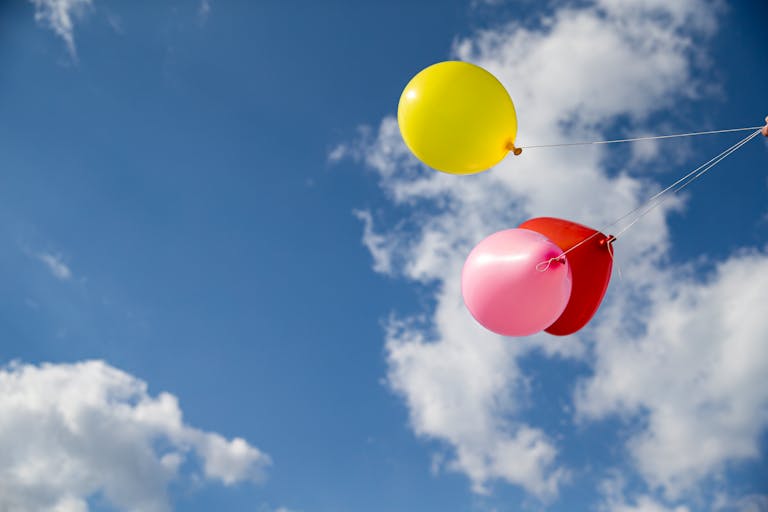 Bright yellow, pink, and red balloons float against a clear blue sky, celebrating summer joy.
