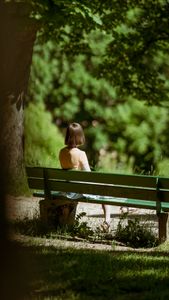 A woman sits peacefully on a green park bench surrounded by lush greenery on a summer day.
